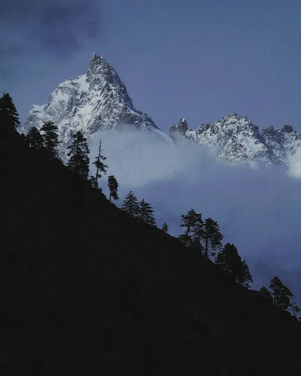 A majestic Himalayan peak emerges from the clouds at dusk. The deep blue and black tones create a moody, powerful scene that reflects the secrets the mountains hold.