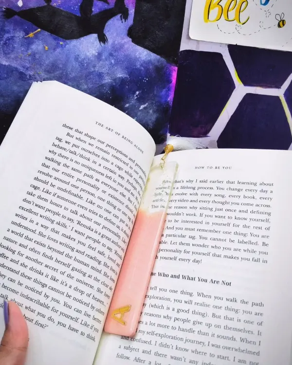 A custom pastel pink bookmark with a gold 'A' initial, shown inside a book. This is how the bookmark looks in use, adding a personal touch to your reading time.