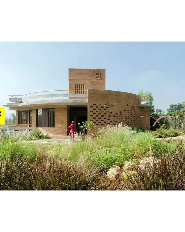 An exterior view of the 'Breathe' residence, showcasing the curved Jaali wall made of compressed earth blocks. The surrounding native grasses and plants are not just decoration but an integral part of the home's ecosystem.