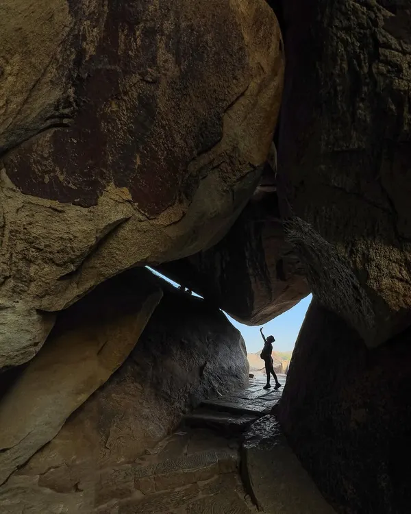 A traveler's silhouette is framed by a natural cave opening, reaching up towards the sky. This photograph captures the sense of adventure and discovery that Hampi's unique landscape inspires.