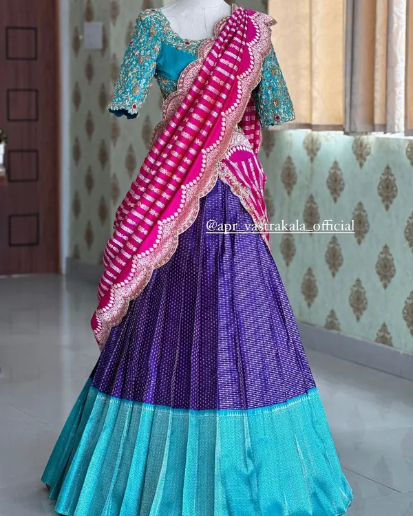 A stunning half saree set displayed on a mannequin. It features a heavily embroidered turquoise blouse, a purple pleated skirt, and a vibrant pink and white striped dupatta with a scalloped border.