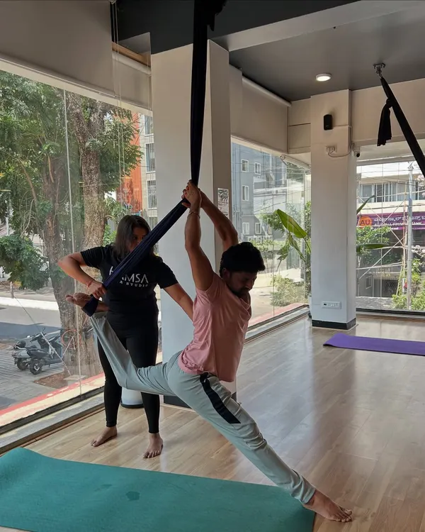 An instructor provides a hands-on assist to a student in a supported split pose during an aerial yoga class.