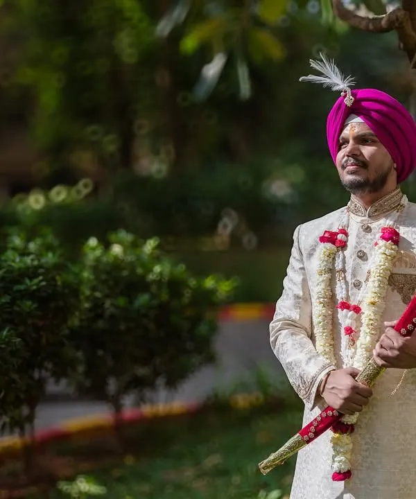 A classic portrait of the groom in his sherwani and magenta turban, holding the ceremonial sword. He looks regal and ready for his wedding.