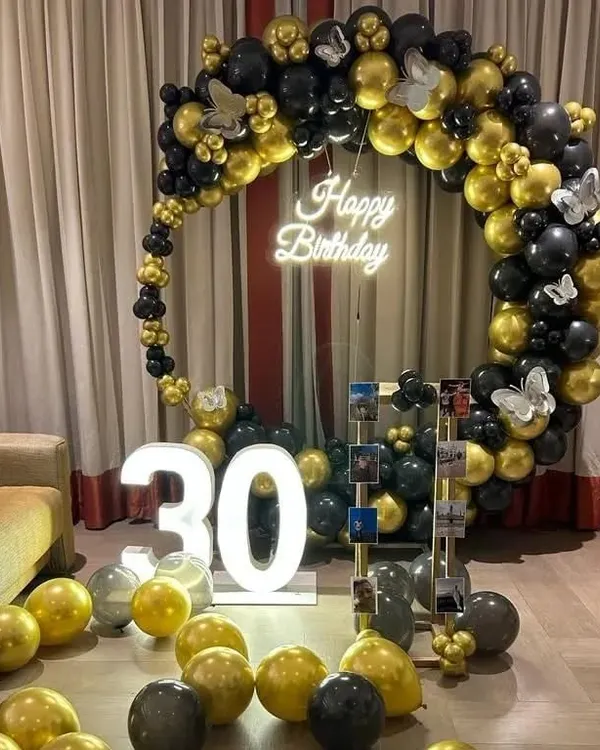 A sophisticated 30th birthday setup with a black and gold circular balloon arch, a 'Happy Birthday' neon sign, and a '30' marquee light. A photo display stand adds a personal touch.