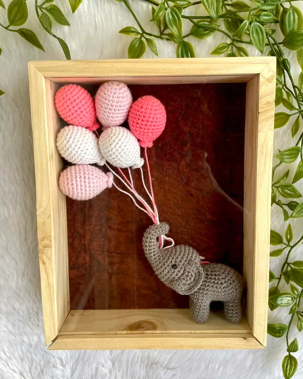 A beautiful nursery wall decor piece featuring a tiny crochet elephant holding a bouquet of pink and white balloons. This 10x8 inch frame comes with unbreakable glass and can be personalized with a name.