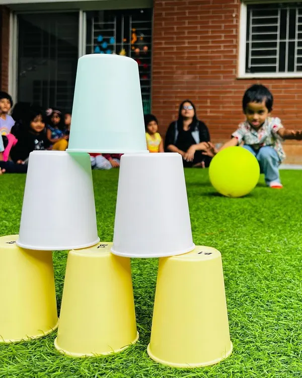 A simple game of cup-stacking bowling helps develop hand-eye coordination and aiming skills. In the background, a toddler gets ready to roll the ball and knock down the tower.