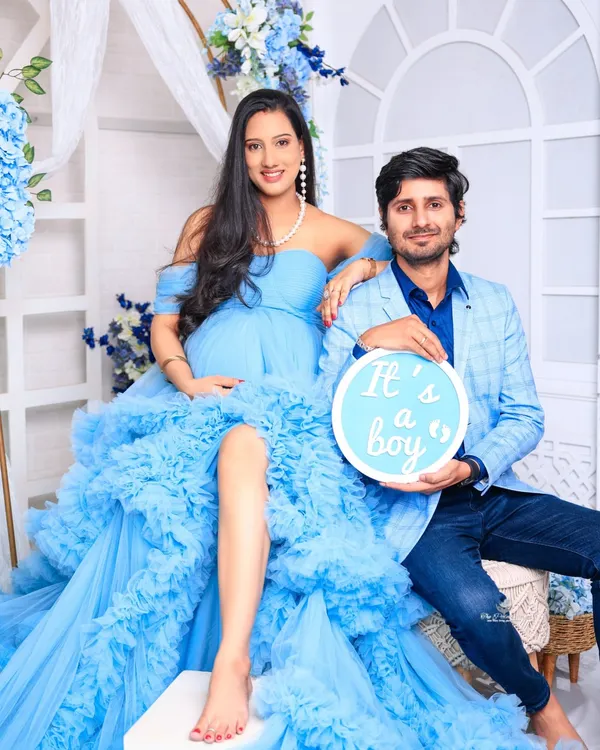 A classic couple's portrait celebrating the upcoming arrival. The mother-to-be is stunning in a ruffled blue gown, while her partner holds an "It's a boy" sign, sharing their joy.
