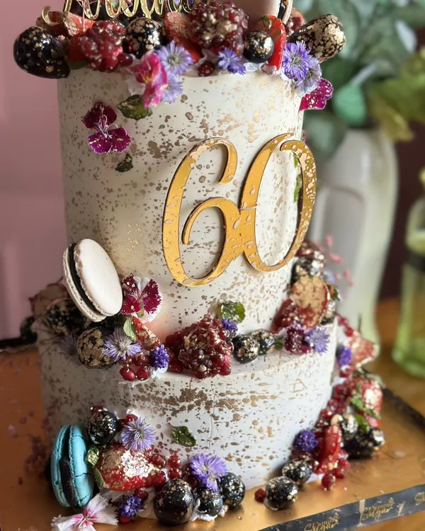 Berry tall, berry extra, berry delicious! This two-tiered cake for a 60th birthday is loaded with fresh berries, macarons, and edible flowers, all dusted with a touch of gold.