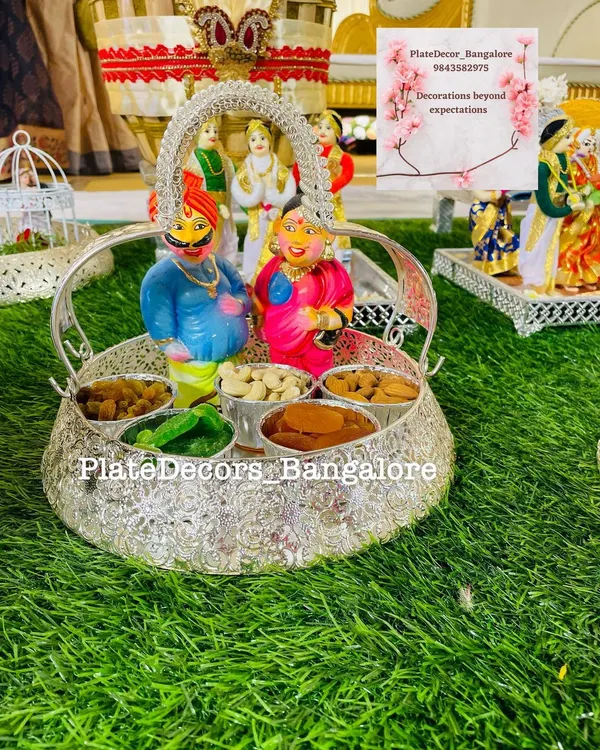 A traditional wedding platter with Marapachi Bommai (couple dolls) made from wood, placed in a silver basket filled with assorted dry fruits.