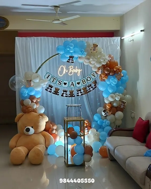 A full view of a teddy bear themed "It's a Boy" baby shower. This cozy home setup features a circular backdrop with a neon sign, a blue, brown, and white balloon arch, and a giant teddy bear.