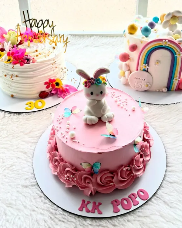 A trio of custom cakes, each with its own unique story. An elegant floral cake for an anniversary, a cute bunny cake for a child, and a rainbow cake for a 5th birthday.