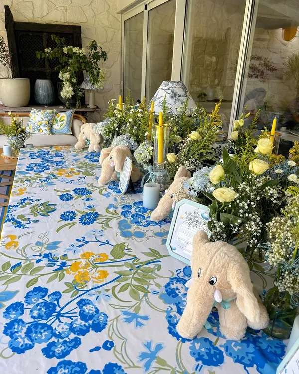 A beautifully set table for a baby's Mundan ceremony, featuring a blue and yellow floral tablecloth, plush elephant toys, and fresh flower arrangements.