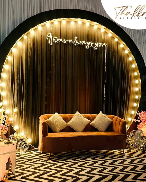 A modern and chic photo booth for a reception. The setup features a circular backdrop with a "It was always you" neon sign, a stylish sofa, and a trendy zig zag floor.