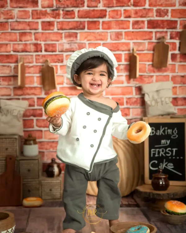 The 'Little Chef' is ready to cook! This happy toddler is holding a toy burger and donut in our custom kitchen set, complete with a brick wall and baking props.