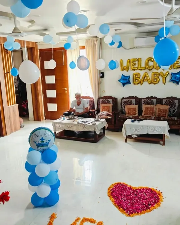 A heartwarming "Welcome Baby" decoration for a home. The setup includes foil letter balloons, balloon bunches hanging from the ceiling, and a flower petal heart on the floor.