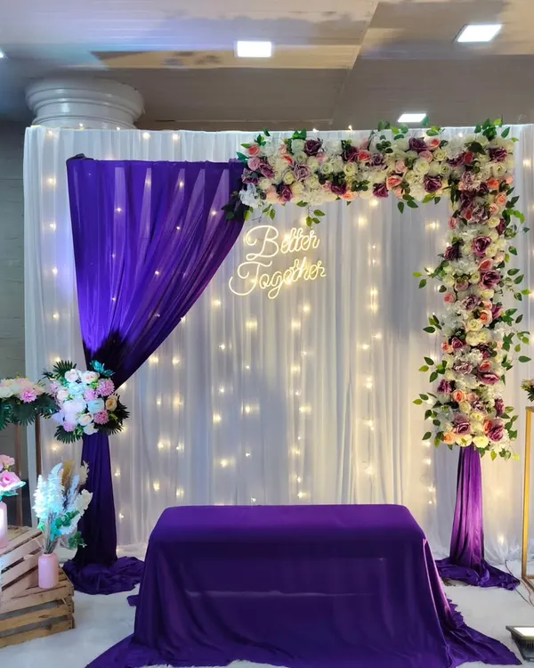 An elegant engagement backdrop with a "Better Together" neon sign, featuring purple and white drapes, a floral arch, and fairy lights.