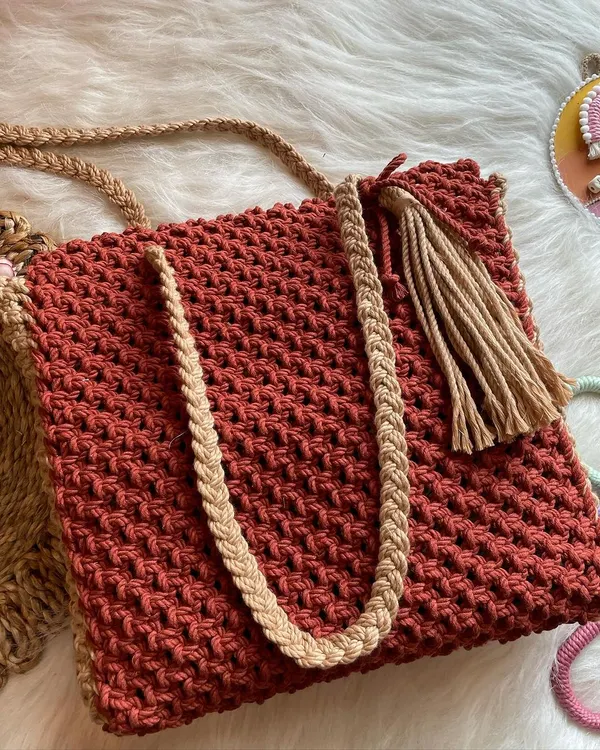 This roomy tote bag in a rich rust color is perfect for everyday use. It features contrasting tan braided straps and a stylish side tassel for a touch of boho flair.