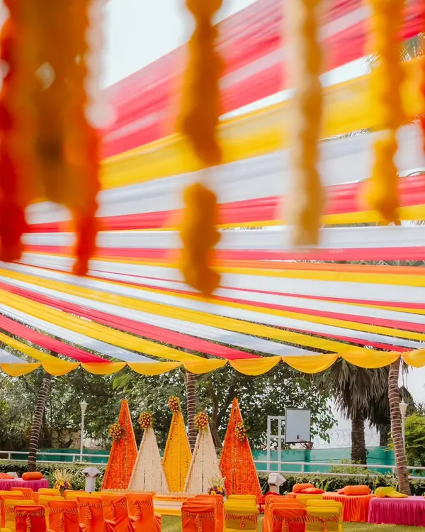A full view of an outdoor Haldi setup, showcasing the vibrant canopy of red, yellow, and white fabric strips. The seating area and floral backdrops are all coordinated to create a bright and joyful environment for the pre-wedding festivities.