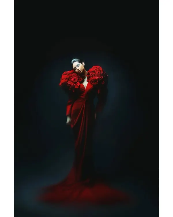 A dramatic portrait from my 'Athena' series for Sigma. The model is styled in a magnificent red gown with floral shoulder details, emerging from a dark background. The lighting is minimal and focused, creating a sense of powerful solitude.