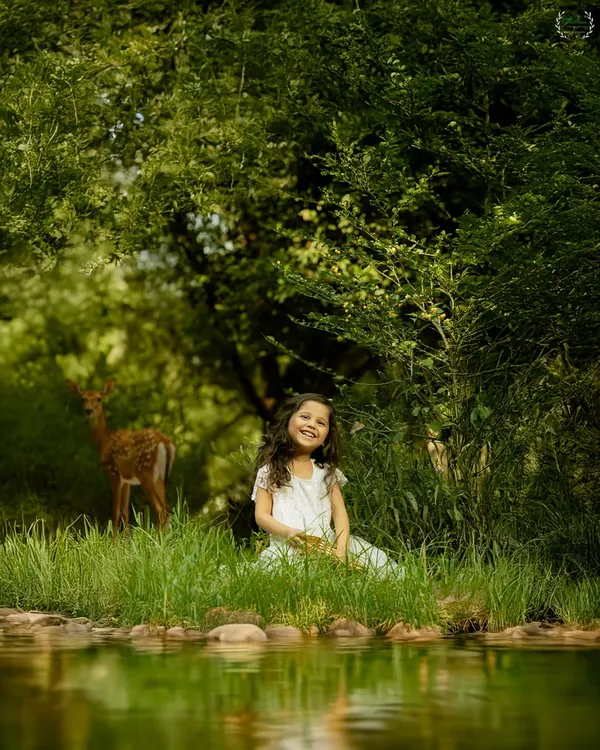 A young girl in a white dress sits by a serene pond, with a deer visible in the lush green background. This magical, storybook-like image showcases the beauty and unpredictability of outdoor photography.