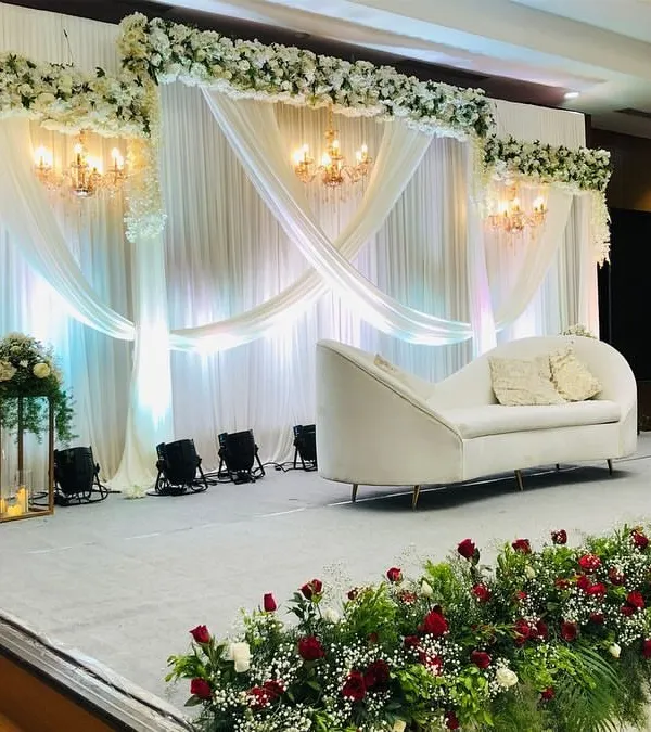 A luxurious engagement stage with flowing white drapes, elegant chandeliers, and a comfortable chaise lounge for the couple. The stage is lined with red and white floral arrangements, adding a touch of romance.