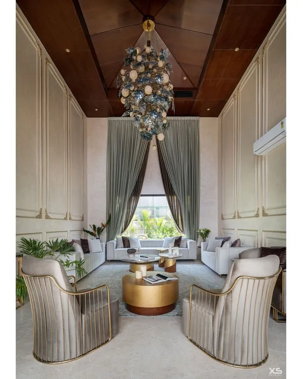 The living room of this luxury villa in Bengaluru features a 22-foot ceiling and a dramatic wooden gable roof. We designed the space with plush, neutral-toned furniture and a massive custom chandelier to create a feeling of royal comfort.