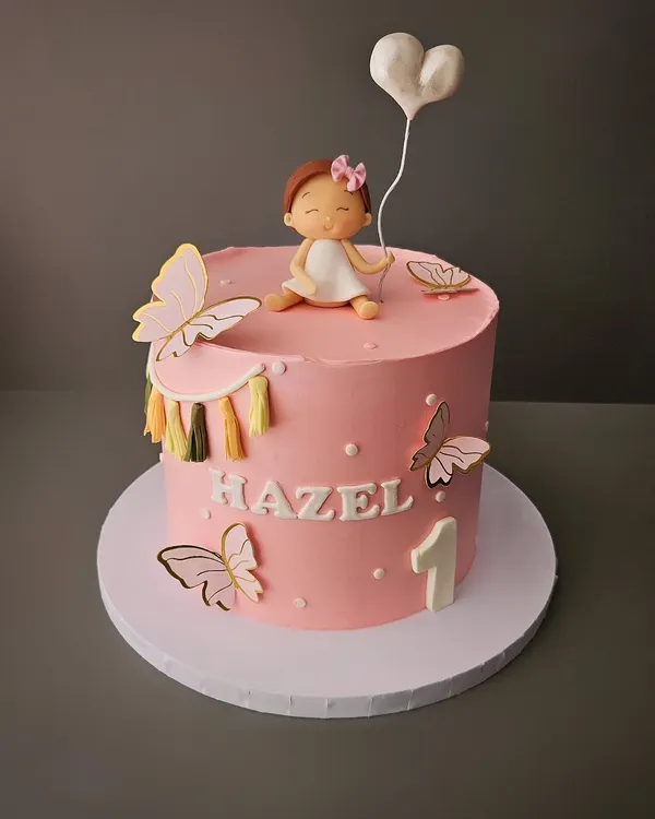 A pretty pink cake for baby Hazel's first birthday. This cake features a cute, hand-sculpted baby figurine holding a heart balloon, surrounded by delicate butterflies.
