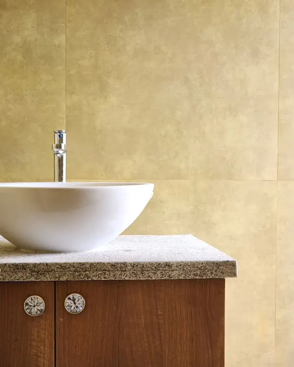 A close-up of a bathroom vanity, featuring a simple white vessel sink on a granite countertop. The brass-toned wall tiles add a warm, metallic sheen, complemented by unique, decorative cabinet knobs.