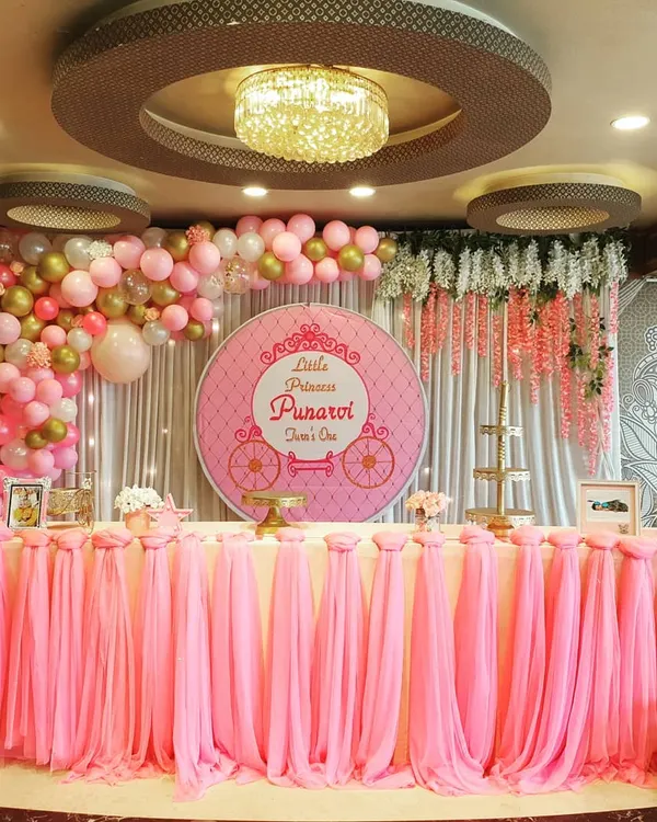 This is the main stage I created for Little Princess Punarvi's first birthday. The setup is defined by a beautifully draped pink table skirt, a custom backdrop with a princess carriage motif, and a cascading balloon arch with pink, white, and gold balloons.
