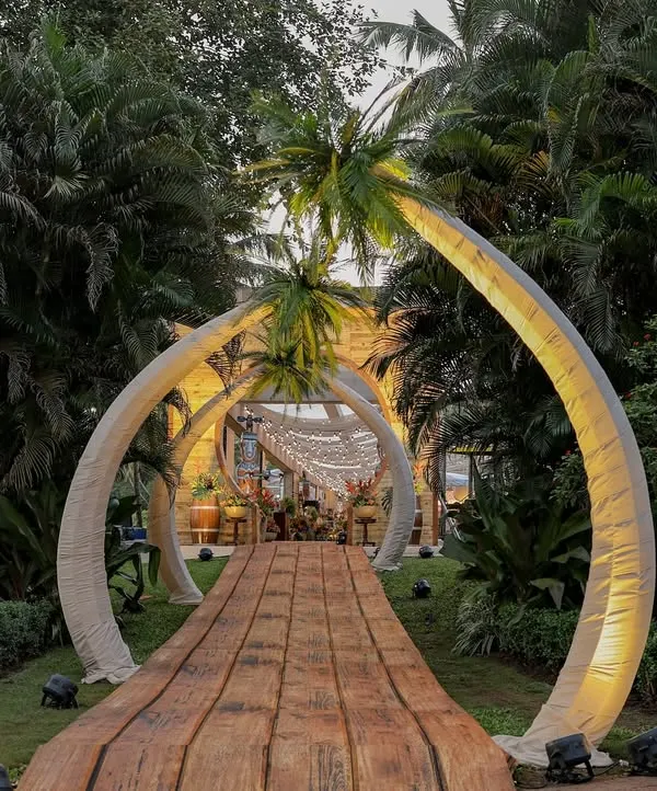 The grand entrance to our 'Bora Bora' themed event. A wooden boardwalk is framed by two massive, curved structures resembling palm trees, creating a dramatic and immersive welcome into our tropical world.