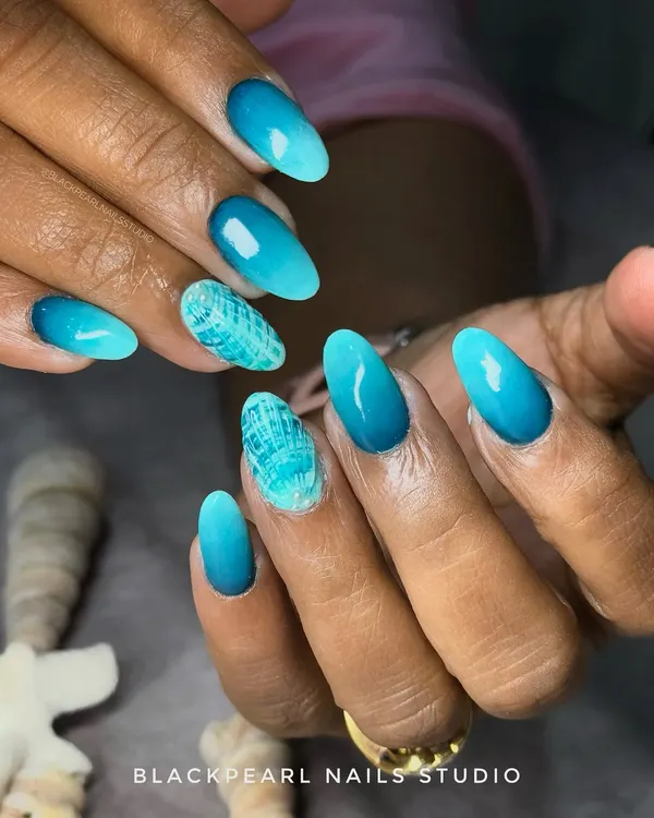 These stunning ocean ombré nails were created for a client's vacation. I blended layers of blue, turquoise, and seafoam shades to capture the look of waves, adding a textured shell effect accent nail for the perfect beach-ready look.