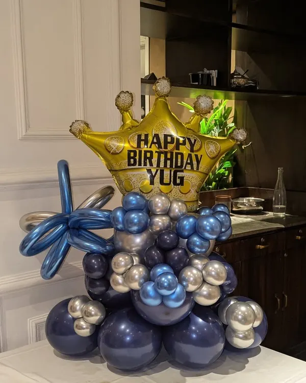 A royal-themed balloon bouquet featuring a large gold crown foil balloon, personalized with the name 'YUG'. The base is made of blue and silver chrome balloons.