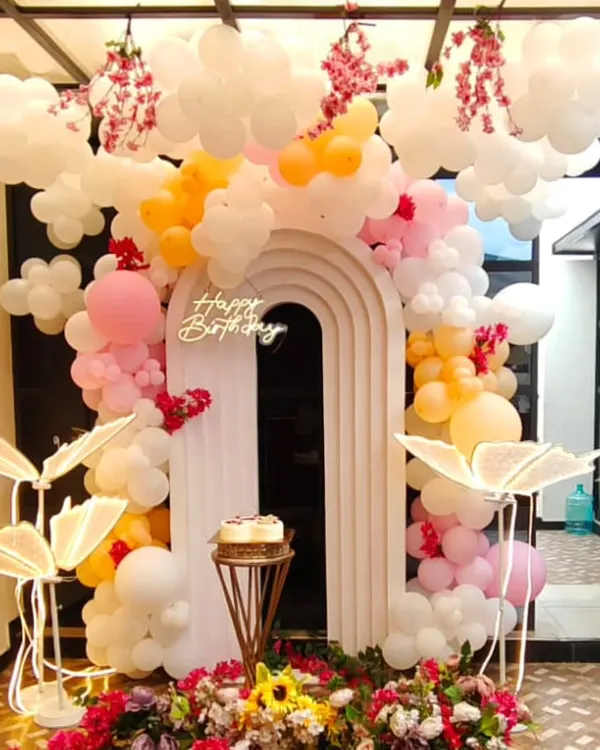 We create dreamlike atmospheres for your special day. This birthday setup features a modern arched backdrop with whimsical balloon clouds in pastel pinks and oranges, a custom neon sign, and beautiful floral arrangements for a truly magical photo corner.