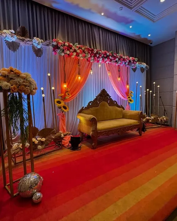 An elegant stage for an anniversary celebration. The decor features a floral runner of pink and white roses, peach-colored drapes, hanging Edison bulbs, and a classic carved sofa.
