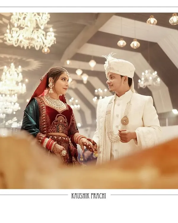 A classic wedding portrait of the couple, Kaushik and Prachi, holding hands after the ceremony. The grand chandeliers and warm lighting of the venue create a regal and timeless backdrop for this beautiful moment.