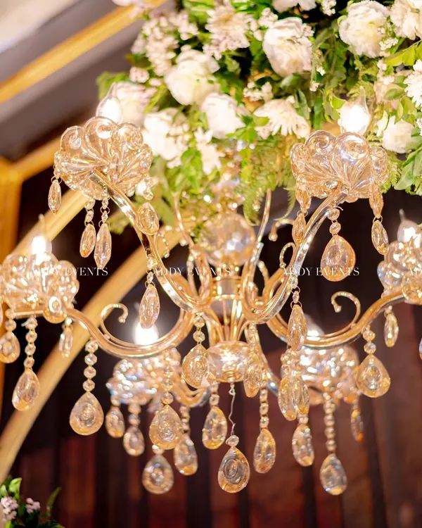 A close-up of pure opulence. A crystal chandelier hangs amidst a lush arrangement of white flowers, a detail that adds glamour and sophistication to any stage or mandap.