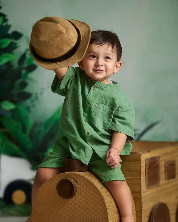 A charming tip of the hat from this little gentleman. This indoor portrait was taken against a jungle-themed backdrop with a wooden train prop, showcasing a fun and creative studio setup.