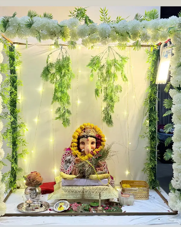 A serene and elegant Ganesh Chaturthi decor for your home. This setup features a simple frame draped with white floral garlands and hanging greenery, illuminated by fairy lights for a divine glow.