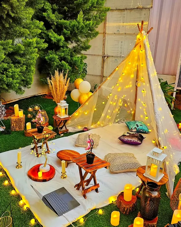 A cozy and romantic nighttime picnic setup. A teepee adorned with fairy lights provides a charming focal point, while the surrounding area is lit with candles and string lights for a magical feel.