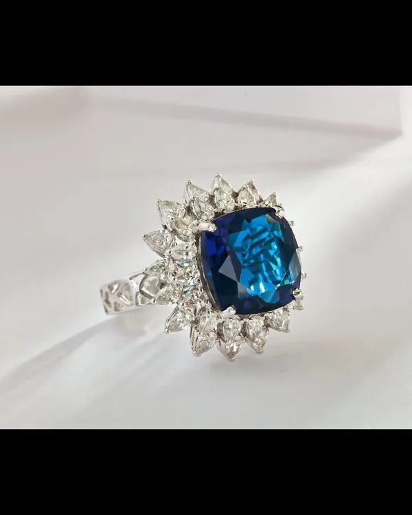 A close-up of a magnificent cushion-cut blue sapphire ring, framed by a brilliant halo of marquise and round diamonds. The studio lighting is carefully controlled to highlight the deep, saturated color of the sapphire and the fire of the surrounding diamonds, making it an ideal shot for a luxury brand's homepage or product detail page.