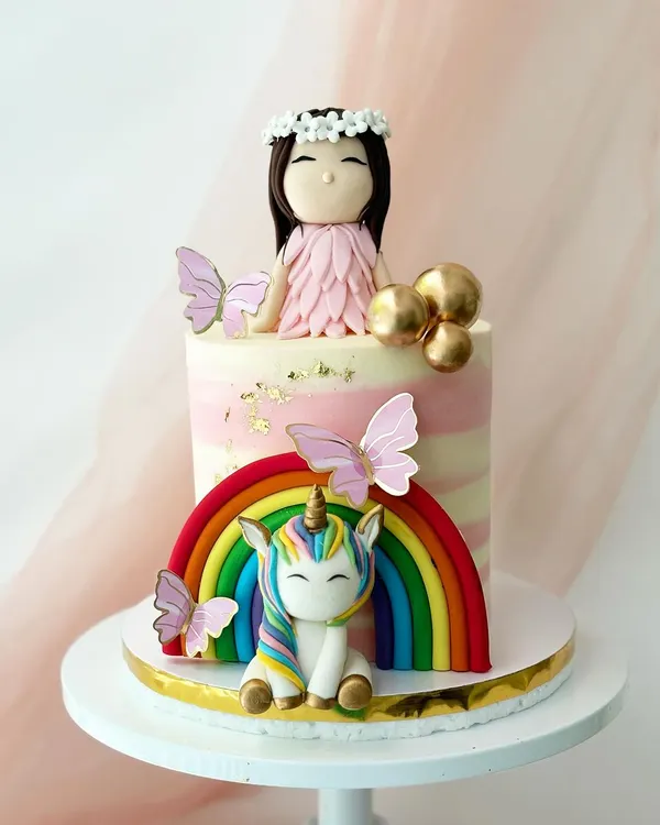 A magical unicorn and fairy theme cake. This design features a rainbow, a cute unicorn, a fairy girl topper, and delicate butterfly accents on a pink striped buttercream base.