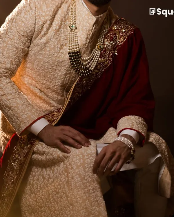 We craft elegant sherwanis that are custom-fitted to make you look and feel exceptional on your special day. This textured beige sherwani, paired with a deep red velvet shawl and pearl accessories, exemplifies the regal aesthetic we deliver for discerning grooms.