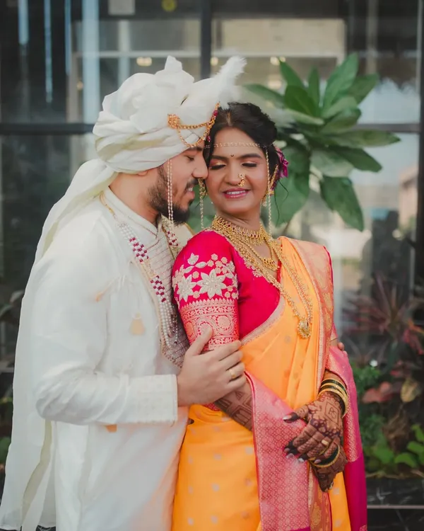 A classic portrait of a Maharashtrian couple, capturing the groom's pride and the bride's gentle smile. The groom is dressed in a traditional white kurta and Pheta, complementing the bride's vibrant yellow and pink Paithani saree.