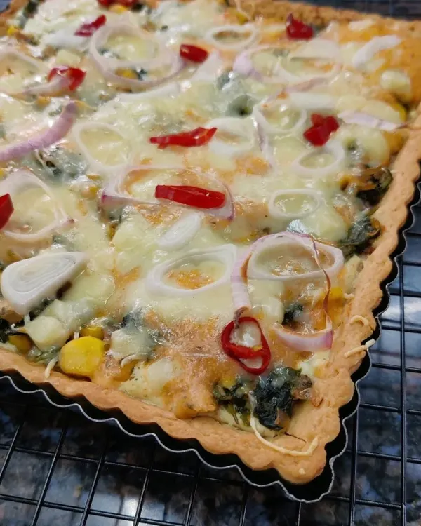 A freshly baked rectangular Corn and Spinach Quiche, with a golden-brown crust, cheesy filling, and topped with rings of onion and red chili.