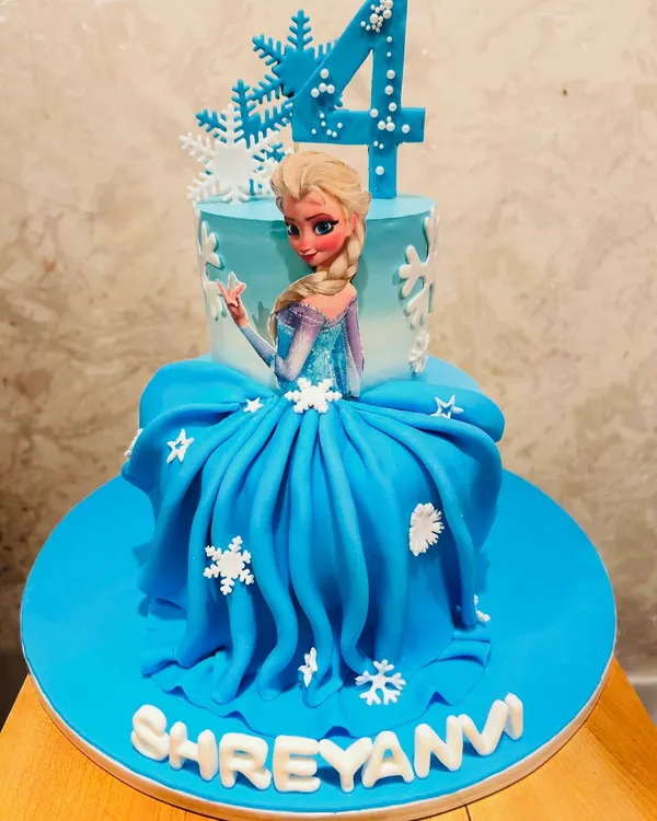 A stunning Frozen-themed doll cake featuring Queen Elsa. The bottom tier is an elaborate gown made of blue fondant, complete with edible snowflakes, making it a showstopper for a 4th birthday.