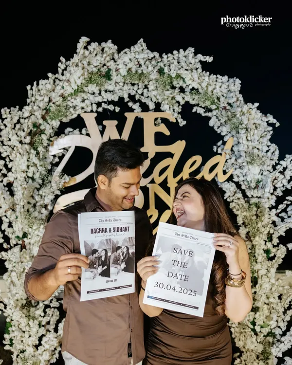 A fun and creative save-the-date photo where a couple holds up custom newspapers announcing their wedding date in front of a floral arch.