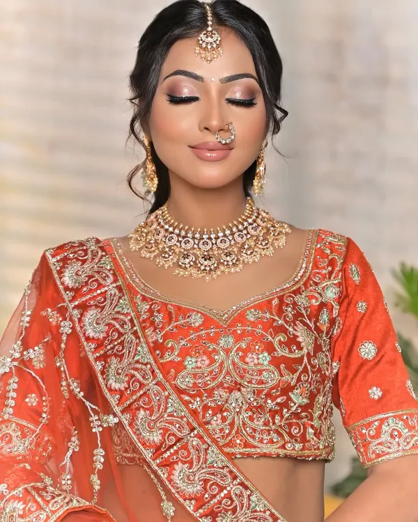 A classic bridal portrait featuring a beautiful Kundan choker with pearl details. This set is paired with a matching maang tikka and a delicate nath, complementing the warm tones of the orange lehenga.