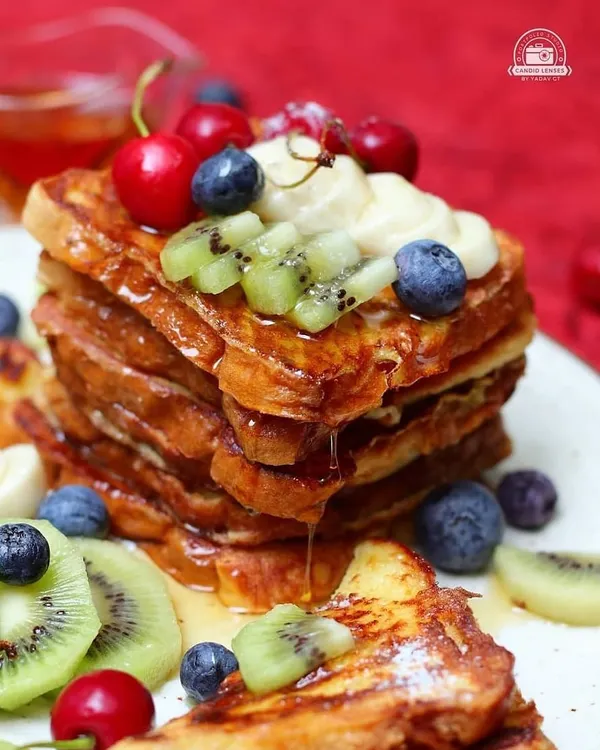 A tall stack of golden French toast is topped with fresh berries, kiwi, and bananas, with syrup dripping down the sides. This close-up food photograph is vibrant, delicious, and expertly styled.