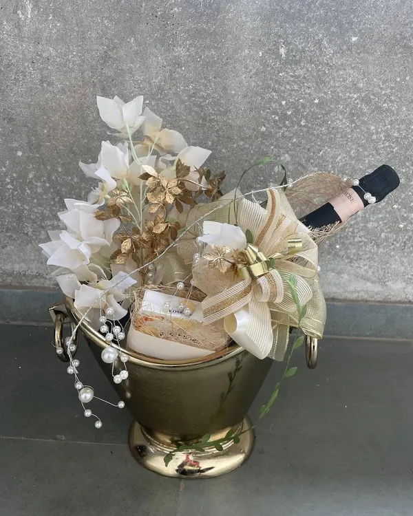 The epitome of luxury bottle gifting. A bottle of champagne is nestled within a golden bucket, lavishly decorated with white and gold florals and cascading pearls, creating a truly show-stopping gift.