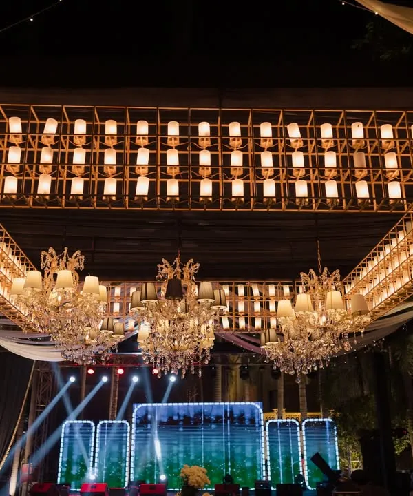 A massive, custom-built chandelier made of hundreds of individual lights serves as a dramatic overhead installation for a Sangeet-style corporate party. Below, the LED stage is ready for performances.